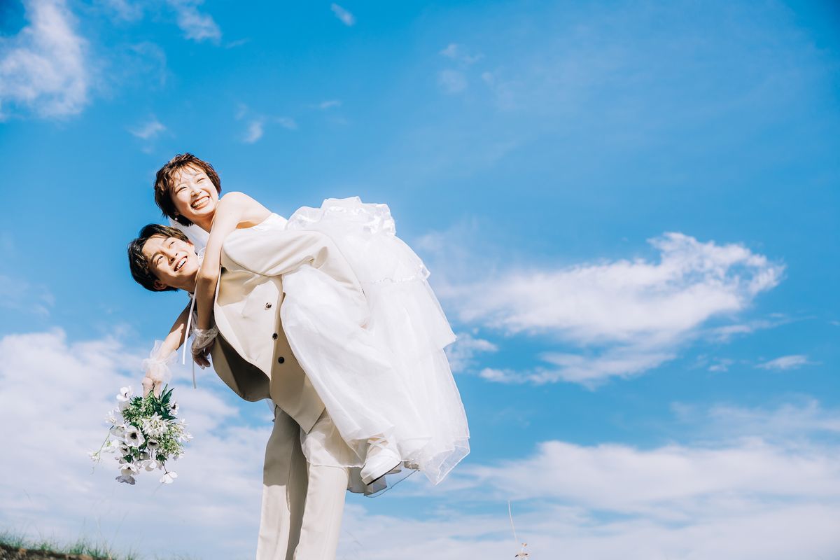 青空の下、花束を持って新郎の背中におぶさり笑う新婦